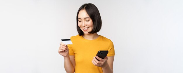 Mooi glimlachend Aziatisch meisje met creditcard en mobiele telefoon die online betaalt op een smartphone die in een gele t-shirt staat op een witte achtergrond