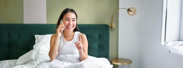 Mooi glimlachend Aziatisch meisje pratend op mobiele telefoon liggend in bed met blij blij gezicht spreken