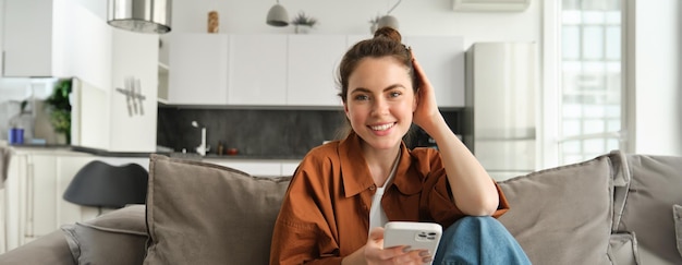 Gratis foto mooi glimlachend vrouwelijk model dat op de bank zit met een smartphone en gelukkig kijkt naar het scrollen van de camera