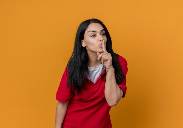 Mooi jong donkerbruin Kaukasisch meisje legt vinger op mond gebaren stil stil teken dat rood overhemd draagt dat op oranje muur wordt geïsoleerd