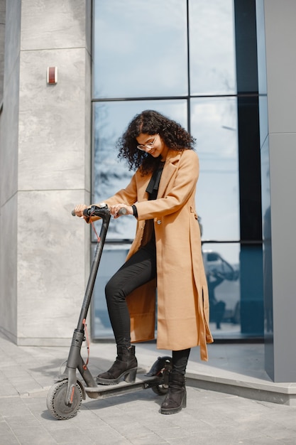 Mooi jong meisje in een bruine jas. Vrouw rijdt op een elektrische scooter.