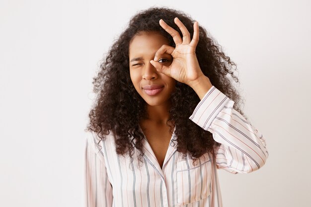 mooi jong mulatmeisje met glanzende gebronsde huid en rommelig krullend kapsel lachend door gat tussen duim en wijsvinger alsof ze een verrekijker gebruikt