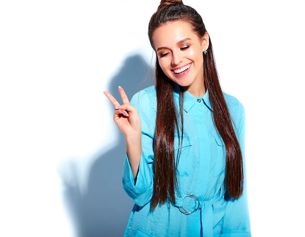 Mooi kaukasisch het glimlachen donkerbruin vrouwenmodel in heldere blauwe de zomer modieuze die kleding op witte achtergrond wordt geïsoleerd. vredesteken tonen