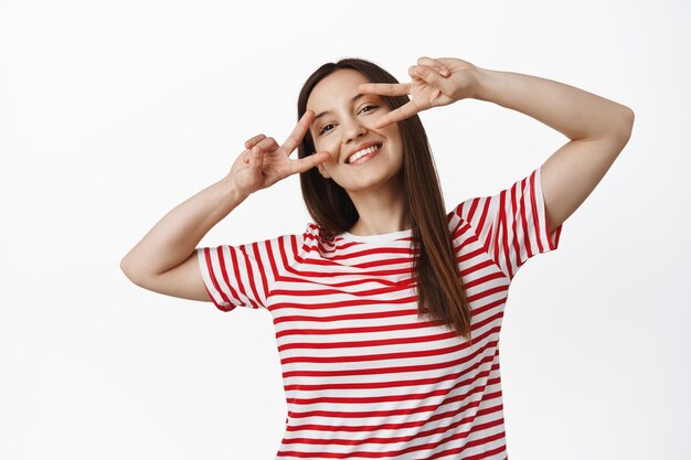 Mooi Kaukasisch meisje vrede v-tekens tonen in de buurt van ogen, glimlachend en op zoek gelukkig, positieve zomerstemming, staande in rood t-shirt tegen witte achtergrond