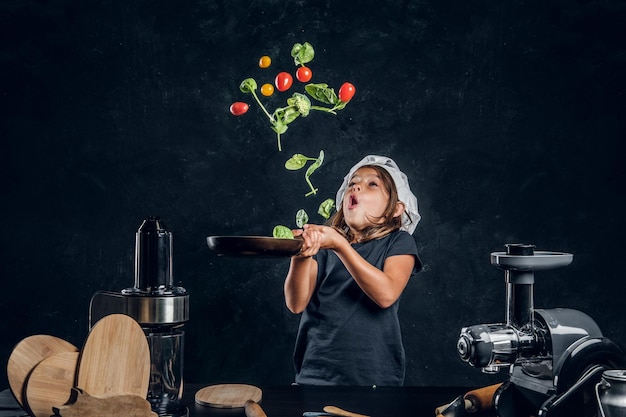 Gratis foto mooi klein meisje gooit groenten op de pan in de donkere fotostudio.