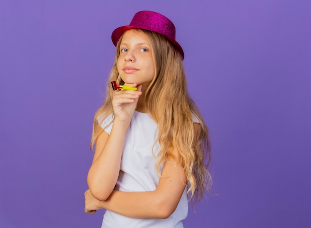 Mooi klein meisje in vakantie hoed bedrijf fluitje kijken camera lachend met blij gezicht, verjaardagsfeestje concept permanent over paarse achtergrond