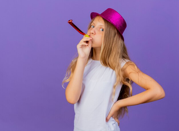Mooi klein meisje in vakantie hoed blaast fluitje kijken camera, verjaardagsfeestje concept permanent over paarse achtergrond