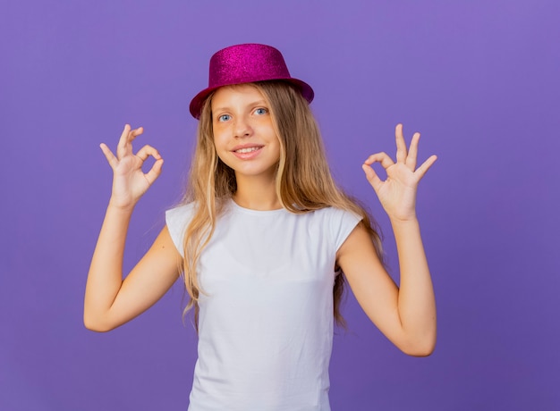 Mooi klein meisje in vakantie hoed kijken camera lachend met blij gezicht weergegeven: ok zingen, verjaardagsfeestje concept permanent over paarse achtergrond