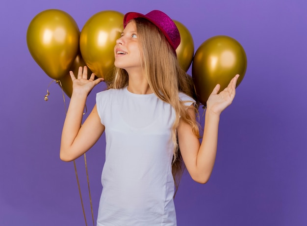 Gratis foto mooi klein meisje in vakantie hoed met bos van ballonnen opzij kijken met blij gezicht, verjaardagsfeestje concept permanent over paarse achtergrond