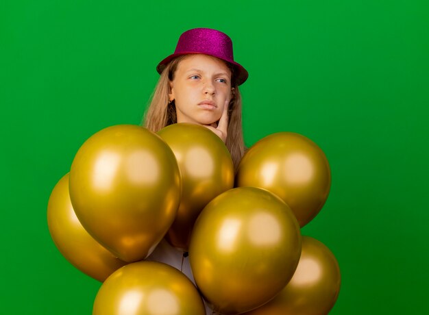 Mooi klein meisje in vakantie hoed met bos van baloons opzij kijken verbaasd, verjaardagsfeestje concept staande over groene achtergrond