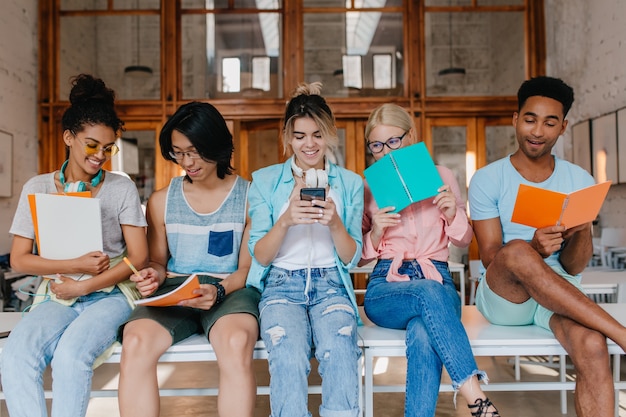 Mooi krullend meisje dat in oortelefoons kijkt wat haar Aziatische vriend toont, die omslagen houdt. Binnenportret van studenten met notitieboekjes die examens in bibliotheek bespreken.