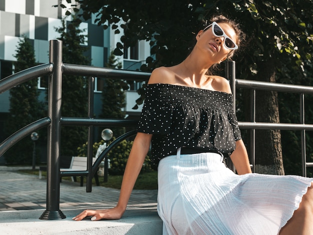 Mooi lachende model gekleed in elegante zomer kleding. sexy zorgeloos meisje zit in de straat. trendy moderne zakenvrouw in zonnebril plezier