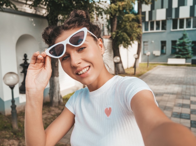 Mooi lachende model met hoorns kapsel gekleed in zomer casual kleding. Sexy zorgeloos meisje poseren in de straat in zonnebril. Selfie zelfportret foto's maken op smartphone