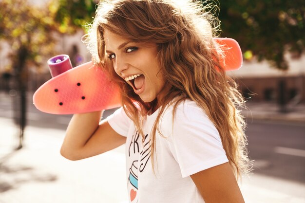 Mooi leuk het glimlachen blond tienermodel zonder make-up in de zomer hipster witte kleren met het roze stuiverskateboard stellen op de straatachtergrond