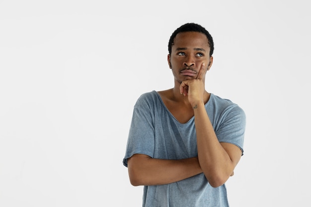 Mooi mannelijk half-lengteportret dat op witte muur wordt geïsoleerd. Jonge emotionele Afro-Amerikaanse man in blauw shirt. Gelaatsuitdrukking, menselijke emoties, advertentieconcept. Nadenken, omhoog kijken.