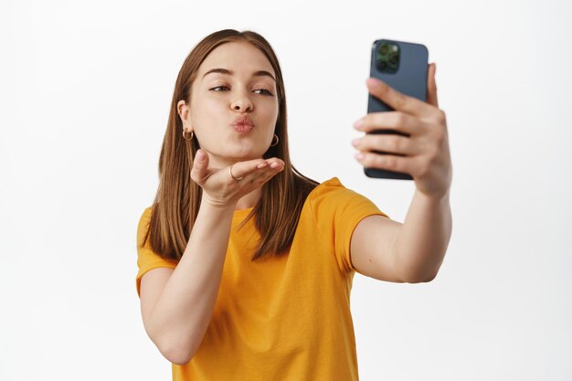 Mooi meisje dat selfie neemt, videochat op mobiele telefoon, luchtkus verzendt op de camera aan de voorkant van de smartphone, flirterig staat in geel t-shirt tegen witte achtergrond