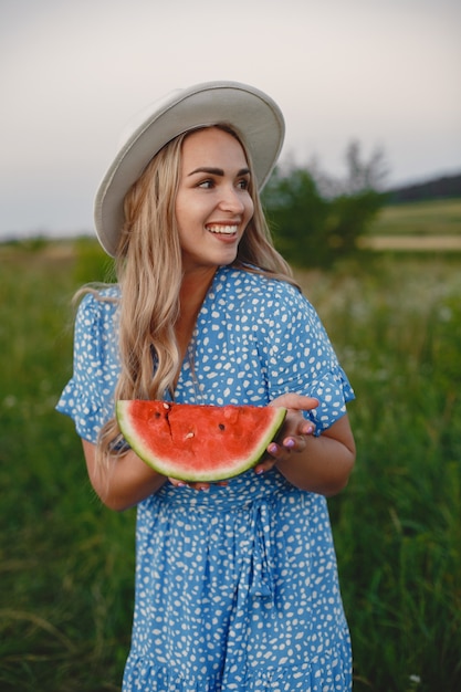 Mooi meisje in een blauwe jurk en hoed. Vrouw in een zomer-veld. Meisje met een watermeloen.