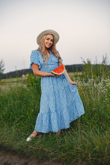 Mooi meisje in een blauwe jurk en hoed. Vrouw in een zomer-veld. Meisje met een watermeloen.