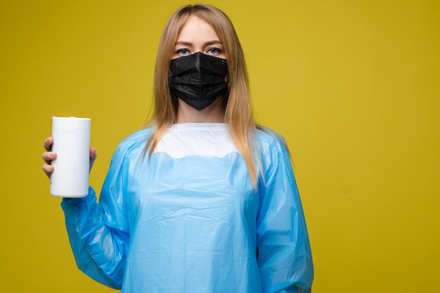 Mooi meisje in een wegwerp medische jurk en met een masker op haar gezicht houdt natte antibacteriële doekjes, portret geïsoleerd op gele achtergrond