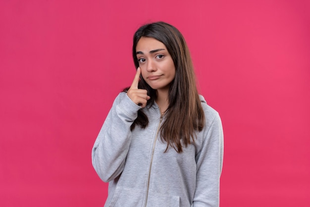 Mooi meisje in grijze hoody camera kijken met droevige uitdrukking wang staande over roze achtergrond aan te raken