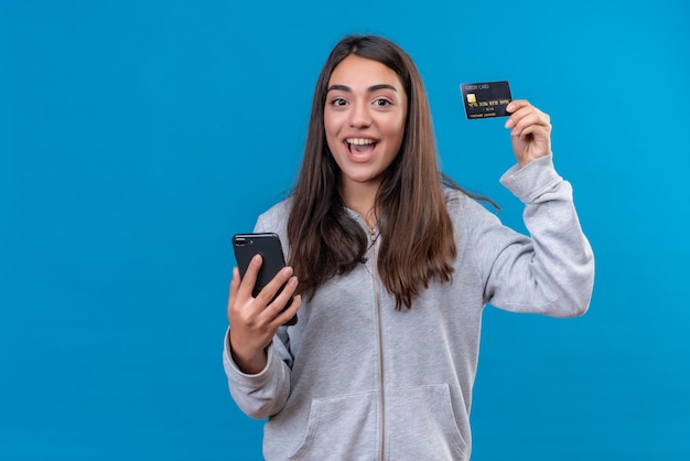 Mooi meisje in grijze hoody camera kijken met glimlach op gezicht en tegelijkertijd telefoon en creditcard staande houden over blauwe achtergrond