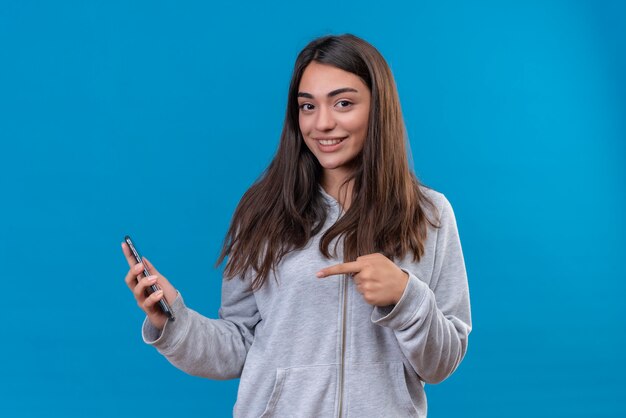Mooi meisje in grijze hoody camera kijken met glimlach op gezicht telefoon houden en wijst naar telefoon staande op blauwe achtergrond