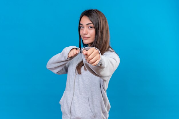 Mooi meisje in grijze hoody camera kijken met winnende emotie en klaar voor strijd staande over blauwe achtergrond