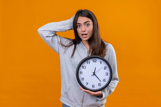 Gratis foto mooi meisje in grijze hoody kijken camera staande met hand op hoofd voor fout en andere hand met klok op haar hand staande over oranje achtergrond