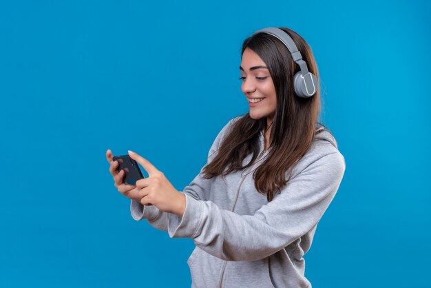 Mooi meisje in grijze hoody met koptelefoon telefoon houden en kijken naar telefoon staande op blauwe achtergrond