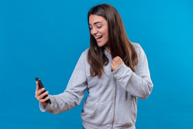 Mooi meisje in grijze hoody telefoon houden en op zoek naar telefoon met glimlach op gezicht staande over blauwe achtergrond