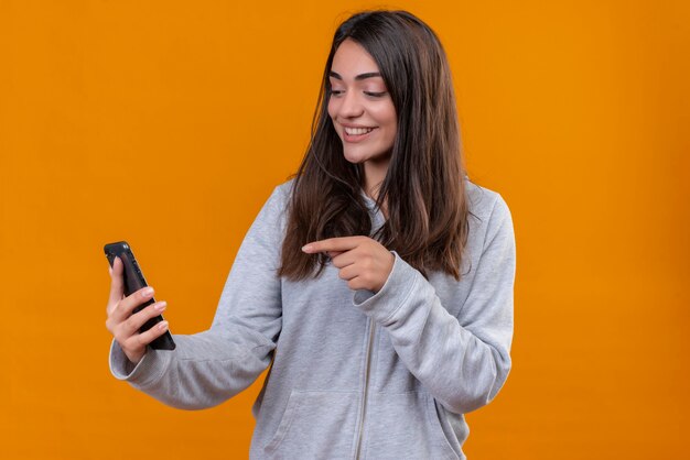 Mooi meisje in grijze hoody telefoon vasthouden en tegelijkertijd naar de telefoon wijzen op zoek naar telefoon met glimlach op gezicht staande over oranje achtergrond