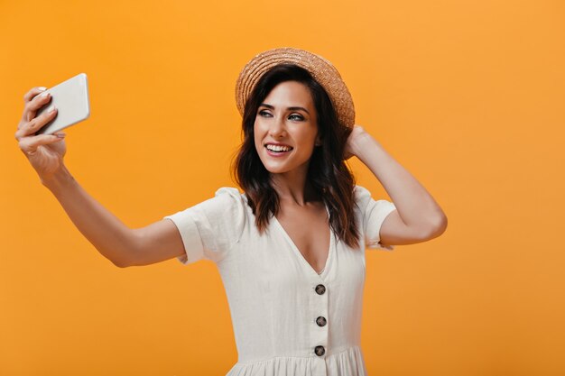 Mooi meisje in hoed maakt gelukkig selfie op een oranje achtergrond. Charmante lachende vrouw in lichte modieuze jurk maakt foto.