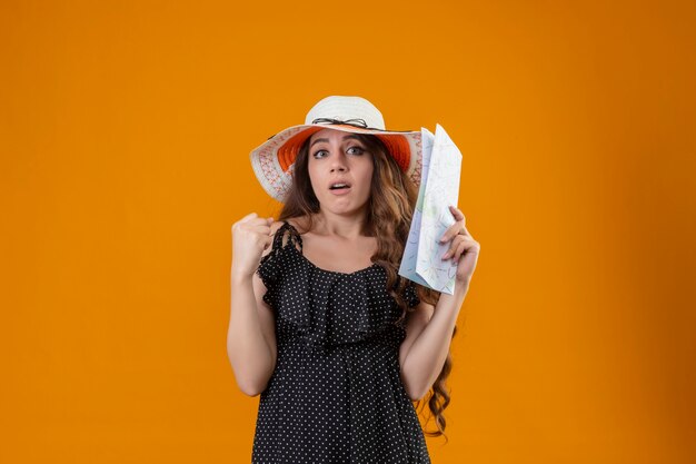 Mooi meisje in jurk in polka dot in zomer hoed bedrijf kaart verhogen vuist verward met sceptische uitdrukking op gezicht staande over gele achtergrond