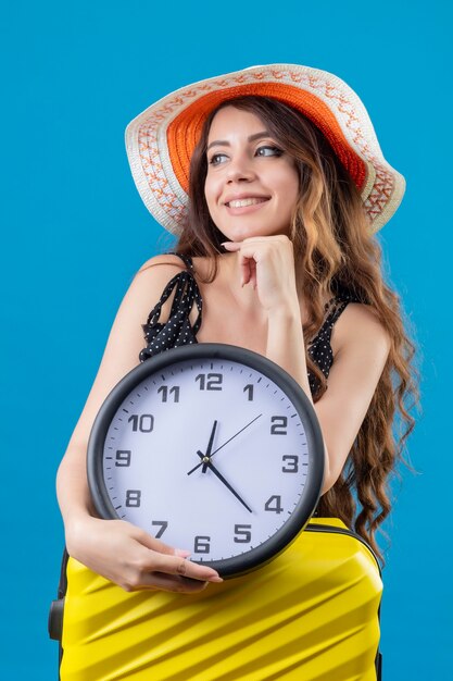Mooi meisje in jurk in polka dot in zomer hoed bedrijf klok staande met koffer wachten met glimlach op gezicht over blauwe achtergrond