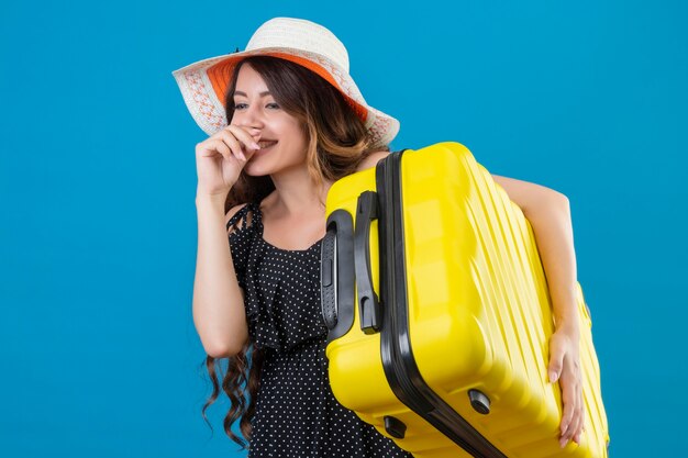 Mooi meisje in jurk in polka dot in zomer hoed bedrijf koffer op zoek positief en gelukkig lachend vrolijk permanent over blauwe achtergrond