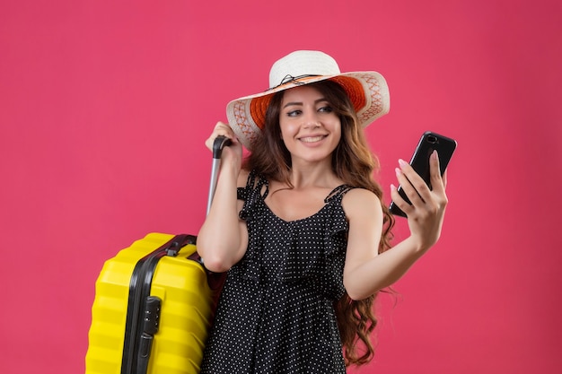 Mooi meisje in jurk in polka dot in zomer hoed permanent met koffer kijken naar scherm van haar mobiele telefoon glimlachend vrolijk op roze achtergrond