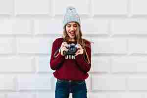 Gratis foto mooi meisje in marsala sweater op grijze muur. ze draagt een gebreide muts, kijkt verbaasd naar de camera in haar handen.
