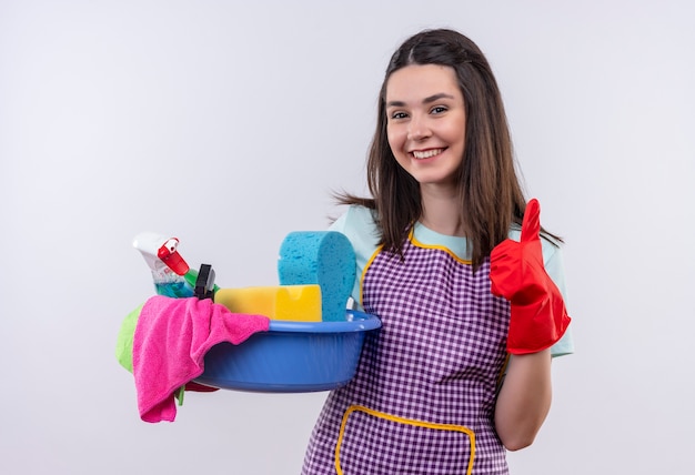 Mooi meisje in schort en rubberen handschoenen bekken met reinigingsgereedschap glimlachend met blij gezicht duimen opdagen te houden