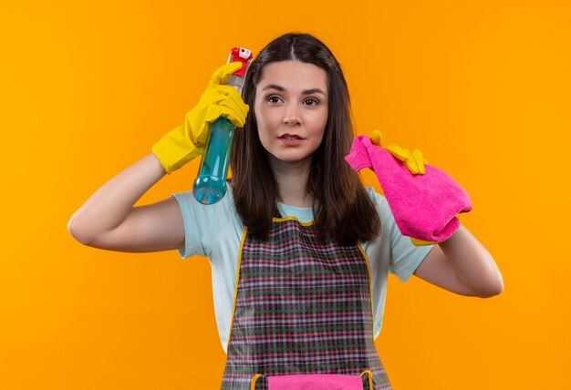 Mooi meisje in schort en rubberen handschoenen met reinigingsspray en tapijt op zoek zelfverzekerd