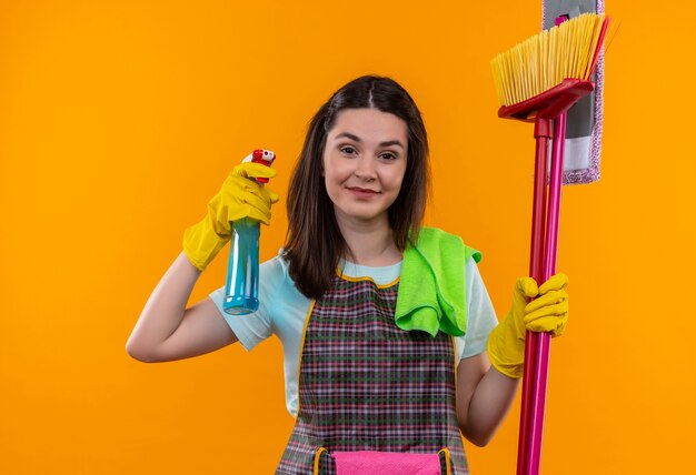 Mooi meisje in schort en rubberen handschoenen met schoonmaak spray en dweilen glimlachend zelfverzekerd, gaan schoonmaken