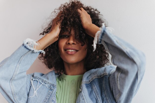 Mooi meisje in spijkerjasje ruches haar op witte achtergrond Portret van Afrikaanse krullende vrouw in groene tee glimlachend op geïsoleerde backdrop