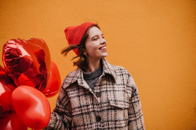 Mooi meisje in stijlvolle hoed lacht oprecht tegen oranje muur Vrouw verheugt zich op Valentijnsdag en poseert met ballonnen in de vorm van harten