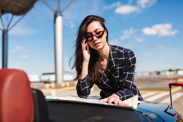 Mooi meisje in zonnebril leunend op zwarte auto zorgvuldig op zoek weg op kaart met luchthaven op achtergrond