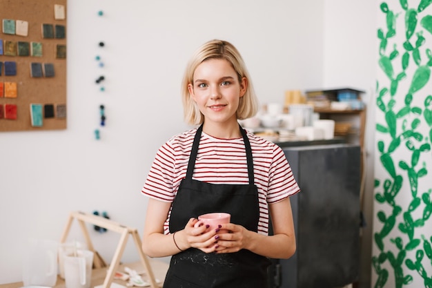 Mooi meisje in zwarte schort met handgemaakte mok in handen en dromerig in de camera kijken naar aardewerk studio