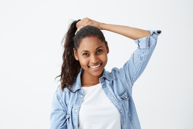 Mooi meisje met een donkere huid die lacht vrolijk haar lange zwarte golvende haar in paardenstaart vastbindt, zich klaarmaakt voordat ze uitgaat met vrienden. Jonge Afro-Amerikaanse vrouwelijke model poseren