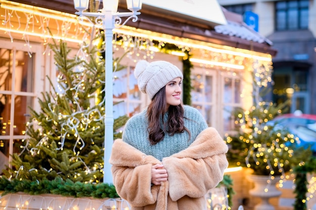 Mooi meisje met een wintermuts en bontjas die in de avondstraat staat, versierd met mooie lichten in de kersttijd
