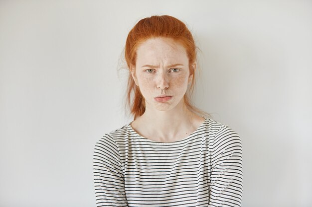 Mooi meisje met gemberhaar, verzameld in paardenstaart die haar lippen pruilt, kijkend met geïrriteerde en beledigde gezichtsuitdrukking terwijl geïsoleerd geïsoleerd