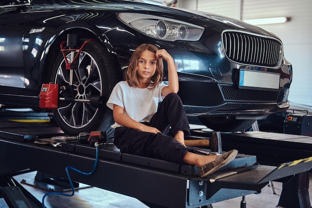 Mooi meisje met gereedschap in de hand zit in de buurt van nieuwe glanzende auto terwijl poseren voor fotograaf.