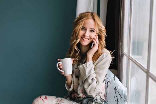 Mooi meisje met golvend haar praten via de telefoon en kopje in de hand houden, zittend op de vensterbank. Kamer met blauwe, turkooizen muur. Een mooie pyjama met bloemen.