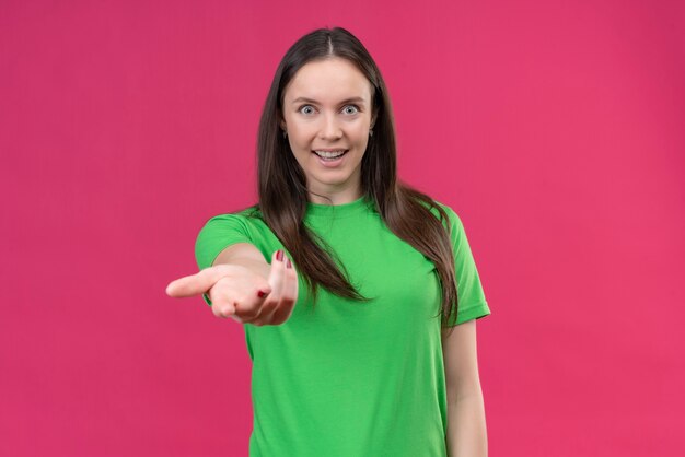 Mooi meisje met groene t-shirt camera kijken met blij gezicht maken kom hier gebaar met hand staande over geïsoleerde roze achtergrond